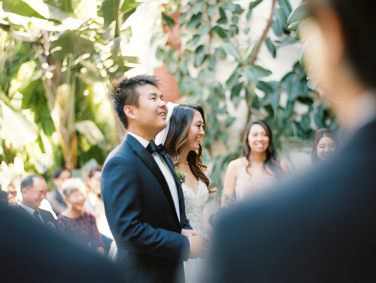 couple who just started wedding ceremony after seeing eachother walk down the aisle at millwick in dtla with palm trees and greenery behind them