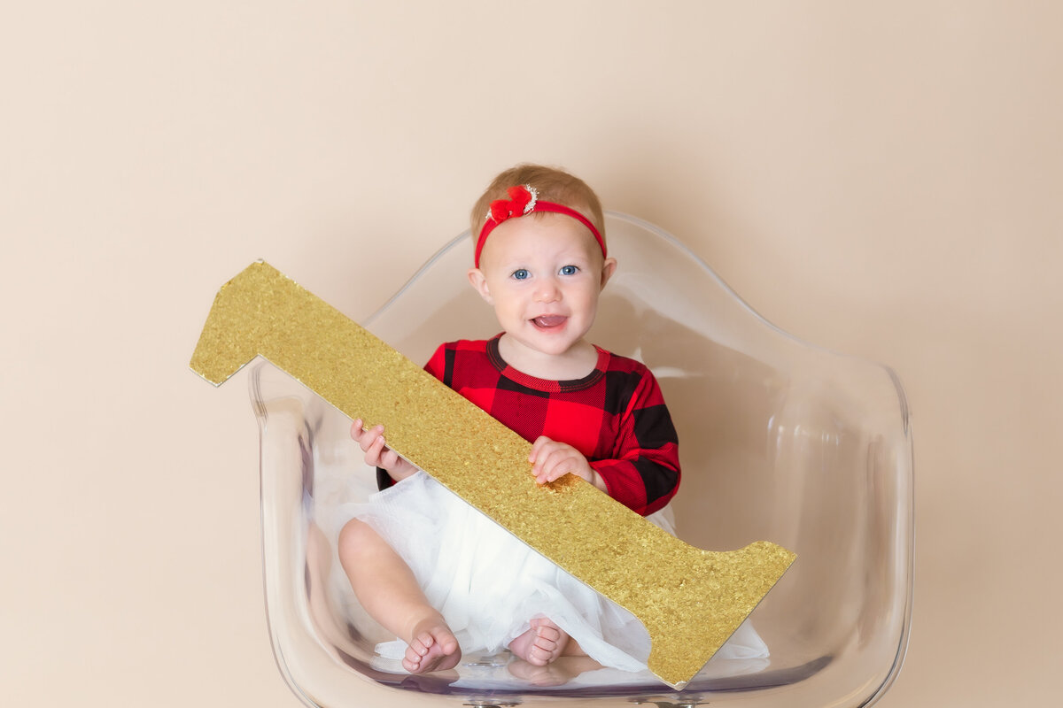 Cake Smash Photographer, a baby smiles on a chair holding a large number one