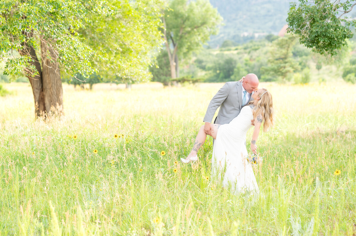 Groom dipping his wife back for intimate moment on their wedding day in open grassy sunflower field