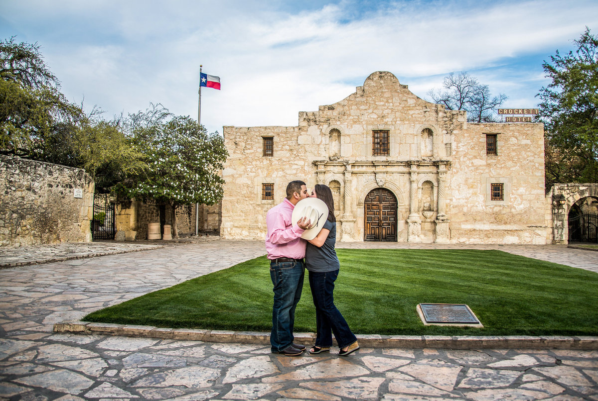 Fiancé and fiancée standing in front of the Alamo in San Antonio hiding their kiss behind a cowboy hat.