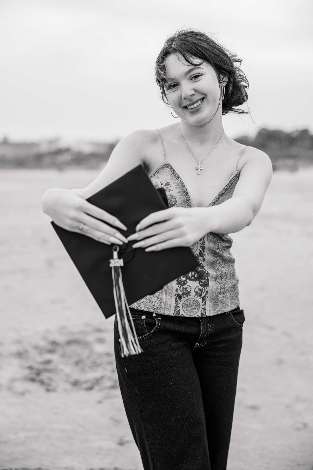 A person is standing on a beach holding a graduation cap with a tassel marked "2021." They are smiling and wearing a patterned sleeveless top and jeans. The background shows a blurred, sandy shore and overcast sky.