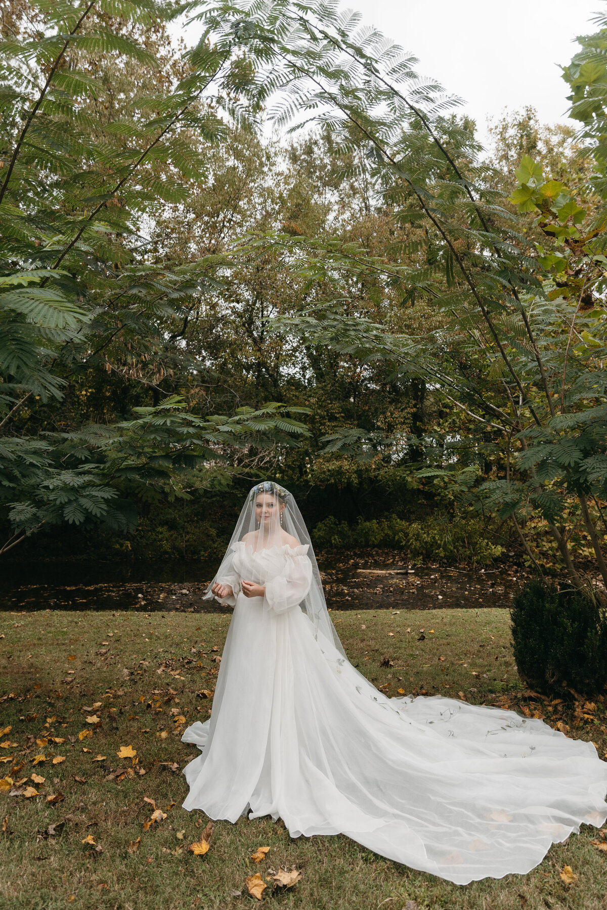 Bride wearing a long flowing cathedral veil with embroidered floral details, standing beneath arching trees in a serene outdoor Arkansas setting—dreamy, whimsical garden wedding portrait.