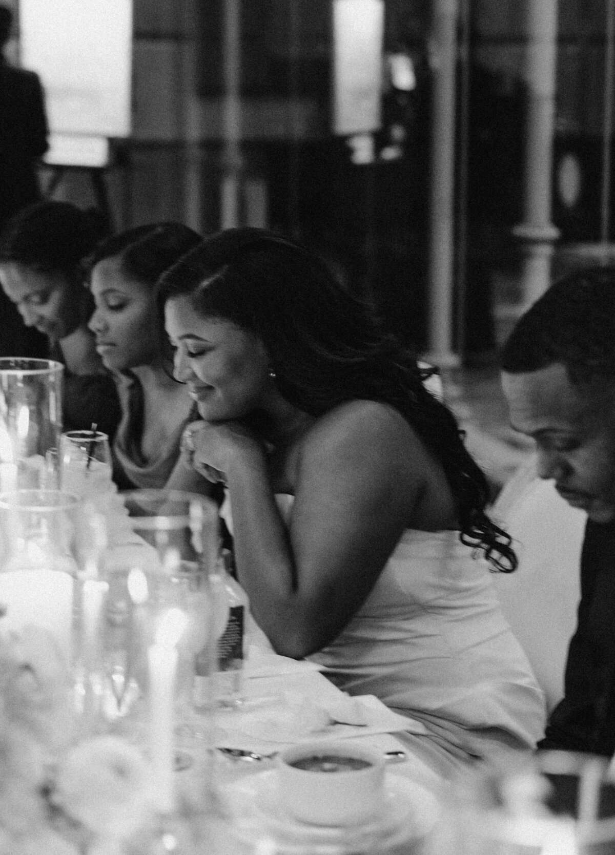 A group of people sits at a dinner table, smiling warmly in a cozy, candlelit setting. The mood is intimate and joyful, with an elegant atmosphere.