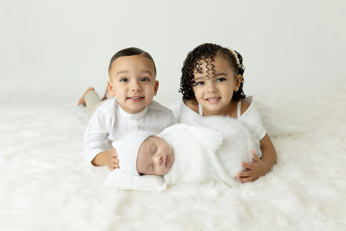 Happy toddler brother and sister lay on a bed holding up their sleeping newborn baby sibling