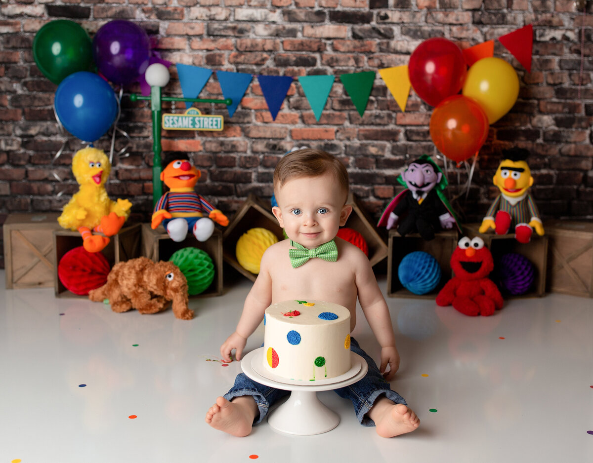 Bright Sesame Street themed cake smash. Baby boy waring jeans and a bowtie sits behind white cake with colorful polkadots. The background is an exposed brick wall, rainbow-colored balloons and pennant with Sesame street characters and street sign.