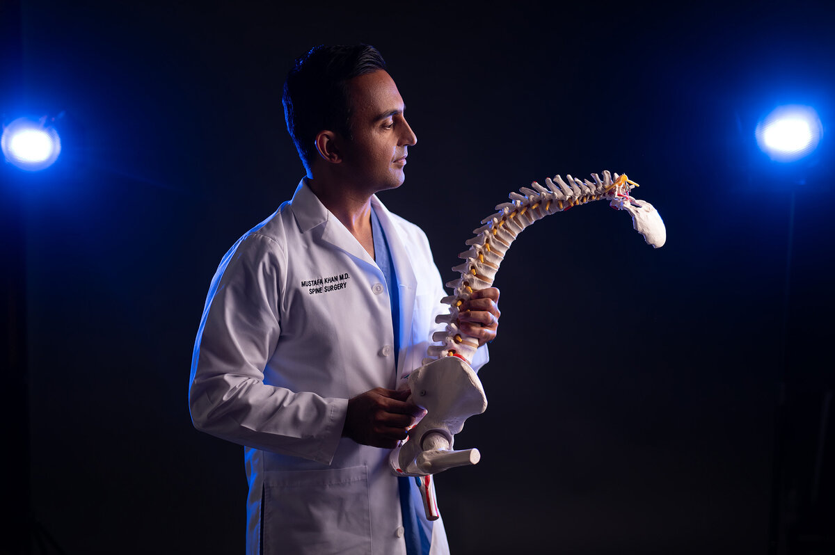 A neurosurgeon in a white coat stands with a spinal model.