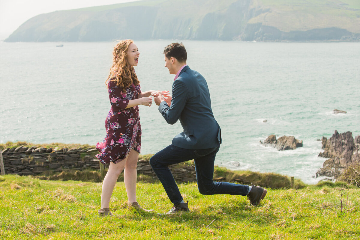 Young man going down on one knee to propose to his girlfriend in a field overlooking the sea