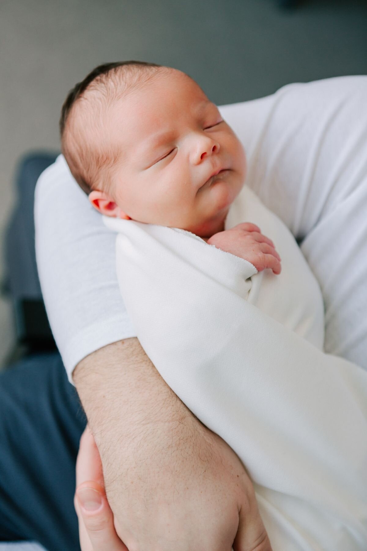 Newborn baby is held in a white blanket, safe in Dad's arms during a newborn photography session with Chelsey Kae Photography