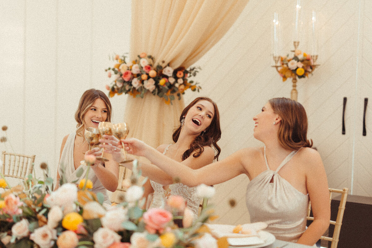 Bride wearing a white wedding gown shares a toast sitting at a table filled with flowers with bridesmaids.