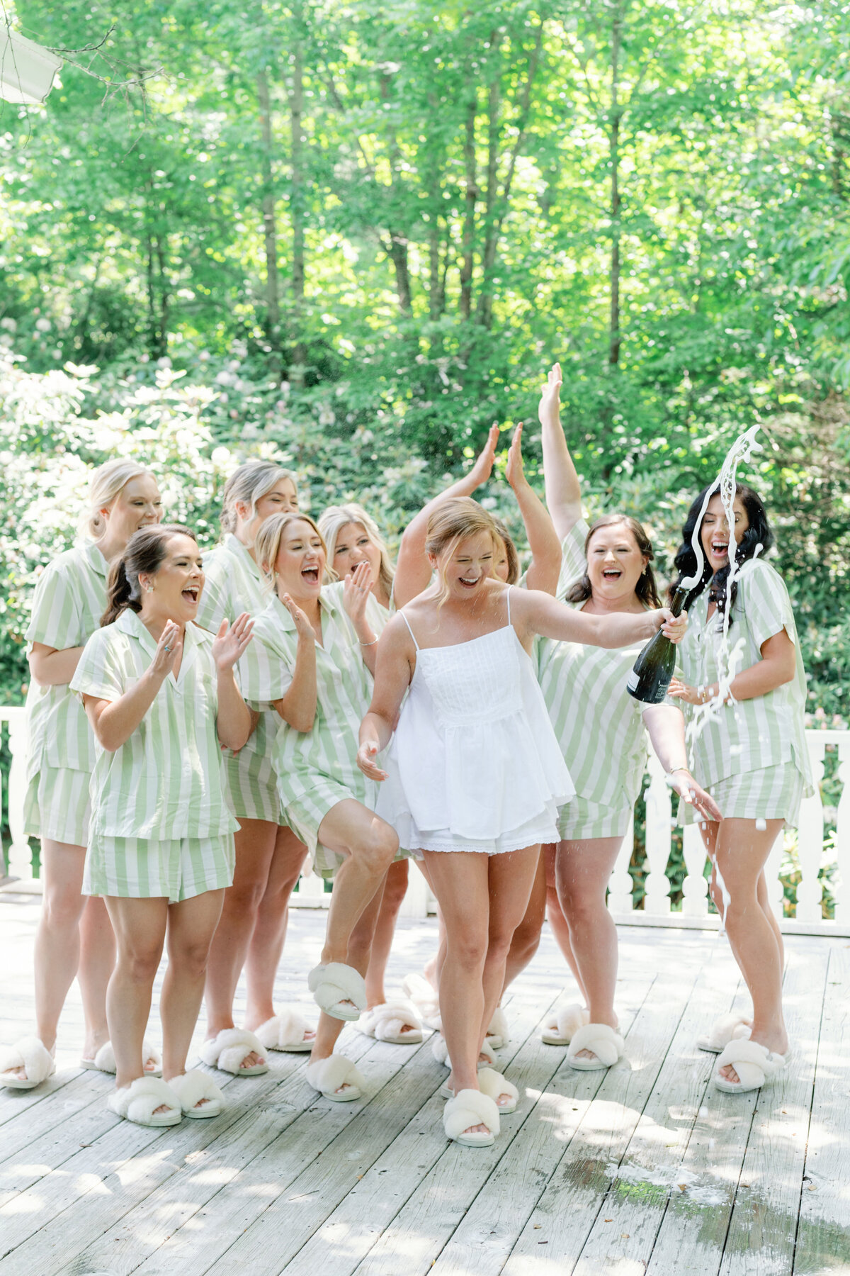 Bride pops champagne in her pjs with bridesmaids. Matching white and green striped wedding pajamas. Summer destination wedding at the Farm at Old Edwards in Highlands, NC.