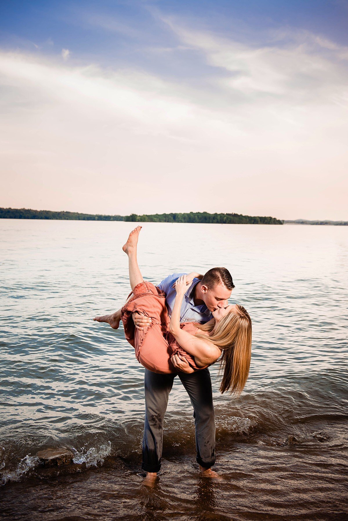 The groom picks the bride up in his arms as he dips her back into a deep kiss. The bride has one bare foot up in the air and her arms around the groom's neck. She is wearing a sleeveless peach sundress. The groom stands barefoot in the waves wearing light gray pants and a light blue shirt.