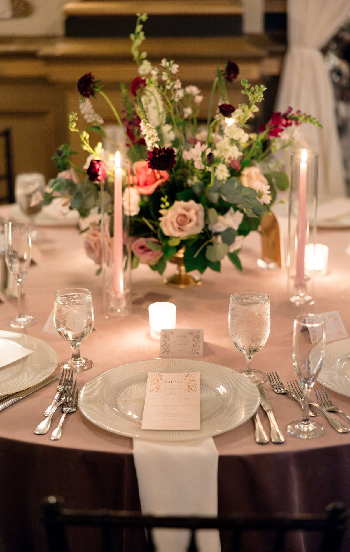 Elegant dining table set for an event, featuring white plates and neatly arranged cutlery. A floral centerpiece with roses and candles adds sophistication. Menus and wine glasses are neatly placed, creating a refined, romantic atmosphere.