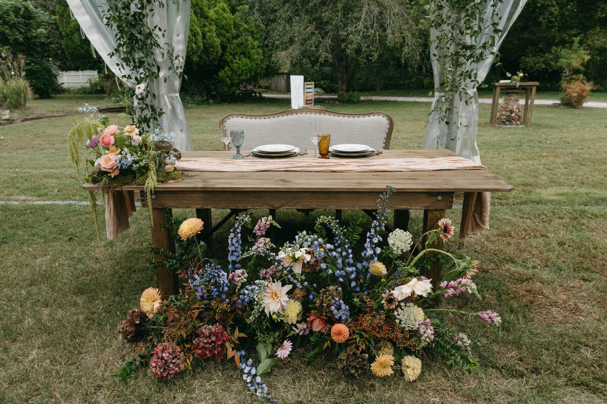Sweetheart table with lush garden-style wedding flowers at The Nest in Northwest Arkansas, featuring whimsical dahlias, delphinium, hydrangea, and autumn-toned roses arranged in a natural, overgrown meadow style by Fleurish Floral Studio. Wooden table with soft linens, vintage-inspired place settings, and a romantic floral ground display under a sailcloth tent.