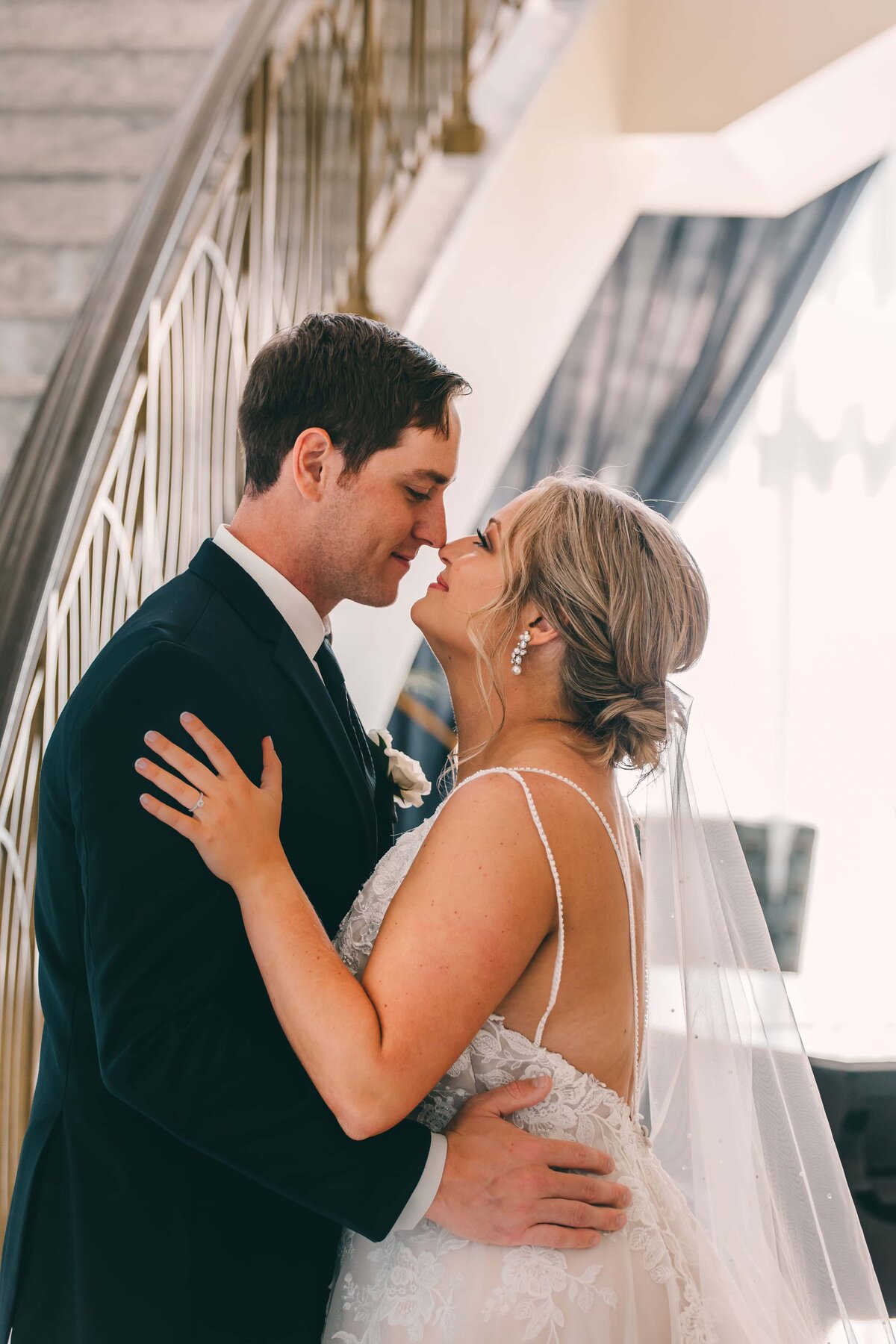 A bride and groom kissing tenderly, with the bride's hand on the groom's cheek, inside a well-lit room at an Iowa wedding with a staircase behind them.