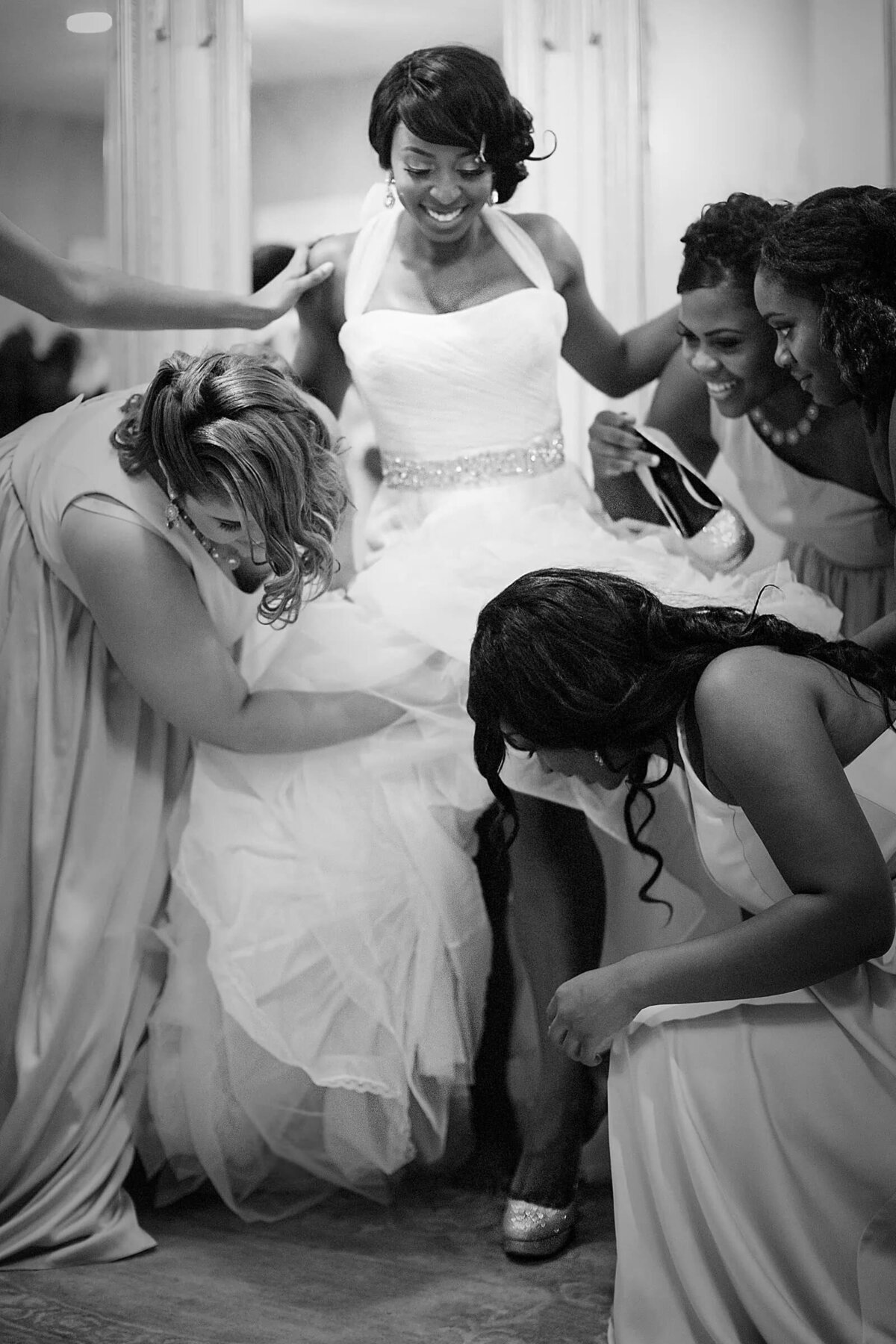 A candid moment in black and white where bridesmaids joyfully help the bride with her dress