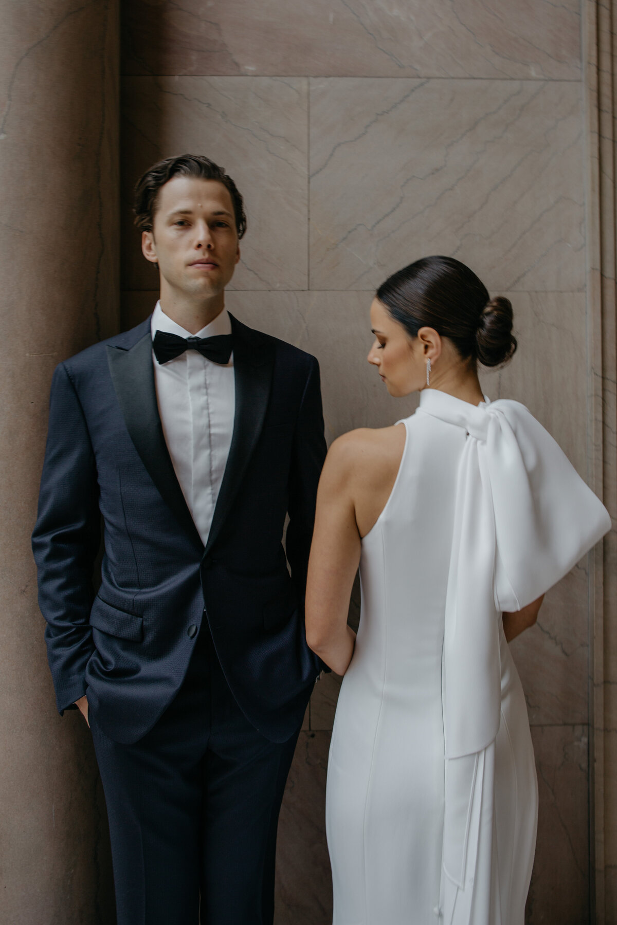 Groom in black tux facing forward with bride turned to side looking down against marble backdrop