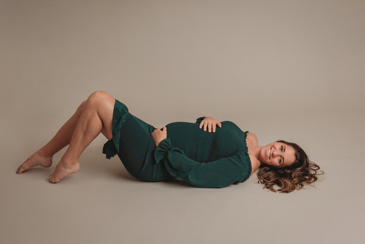 35 week pregnant woman in green dress laying on back smiling at camera with knees up on light gray background