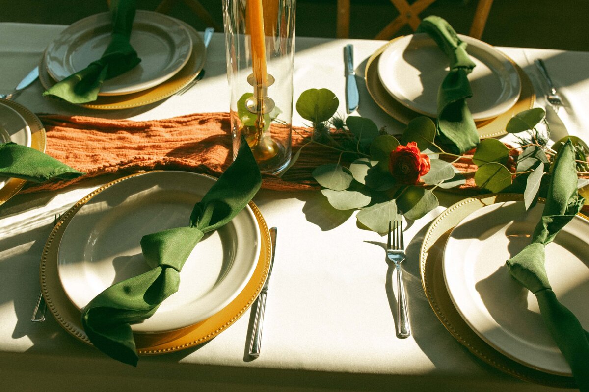 Elegant wedding table setting featuring white plates with gold rims, green napkins, and a centerpiece of greenery and a red flower, illuminated by natural sunlight.