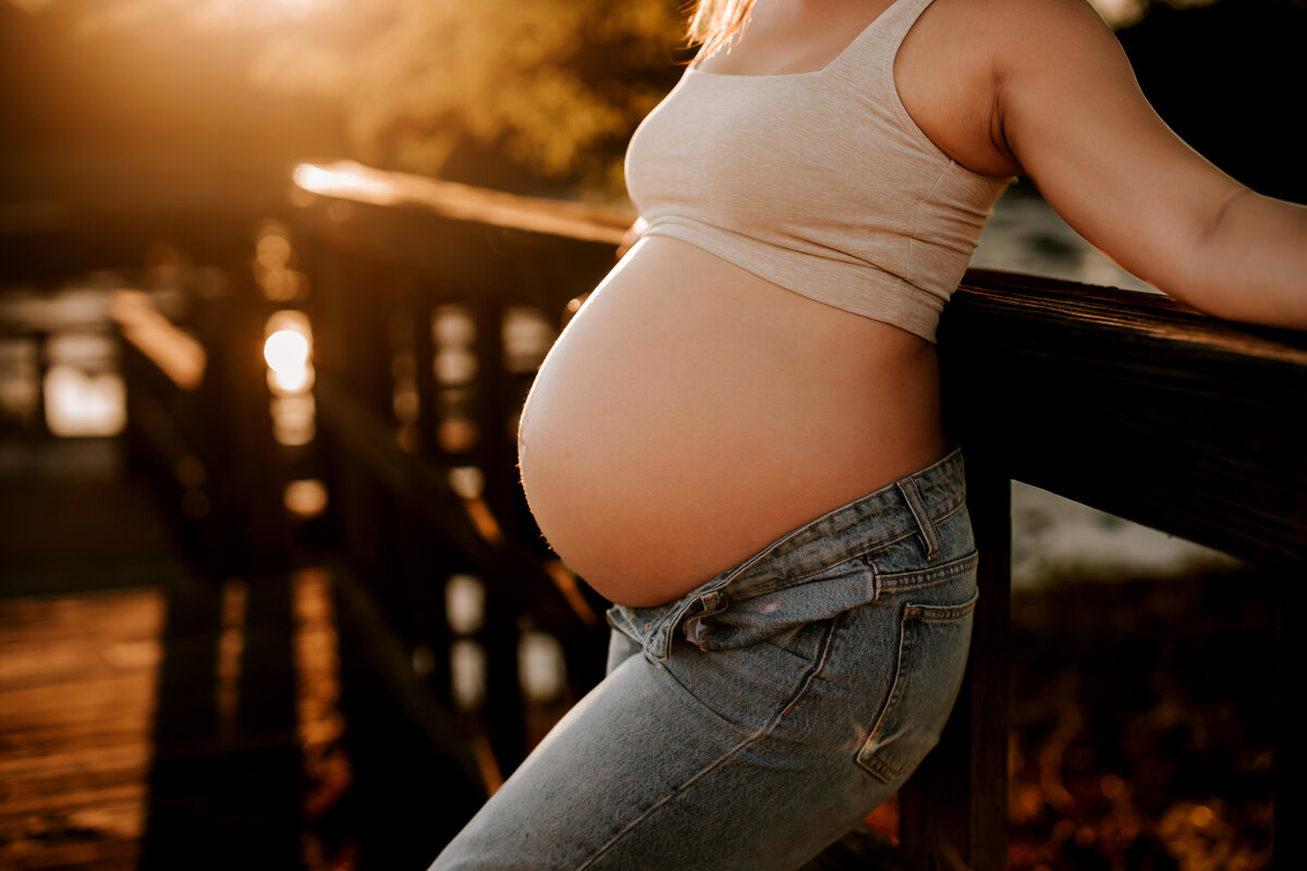 pregnant mom shows off bare belly in jeans while bright sun behind her on boat dock