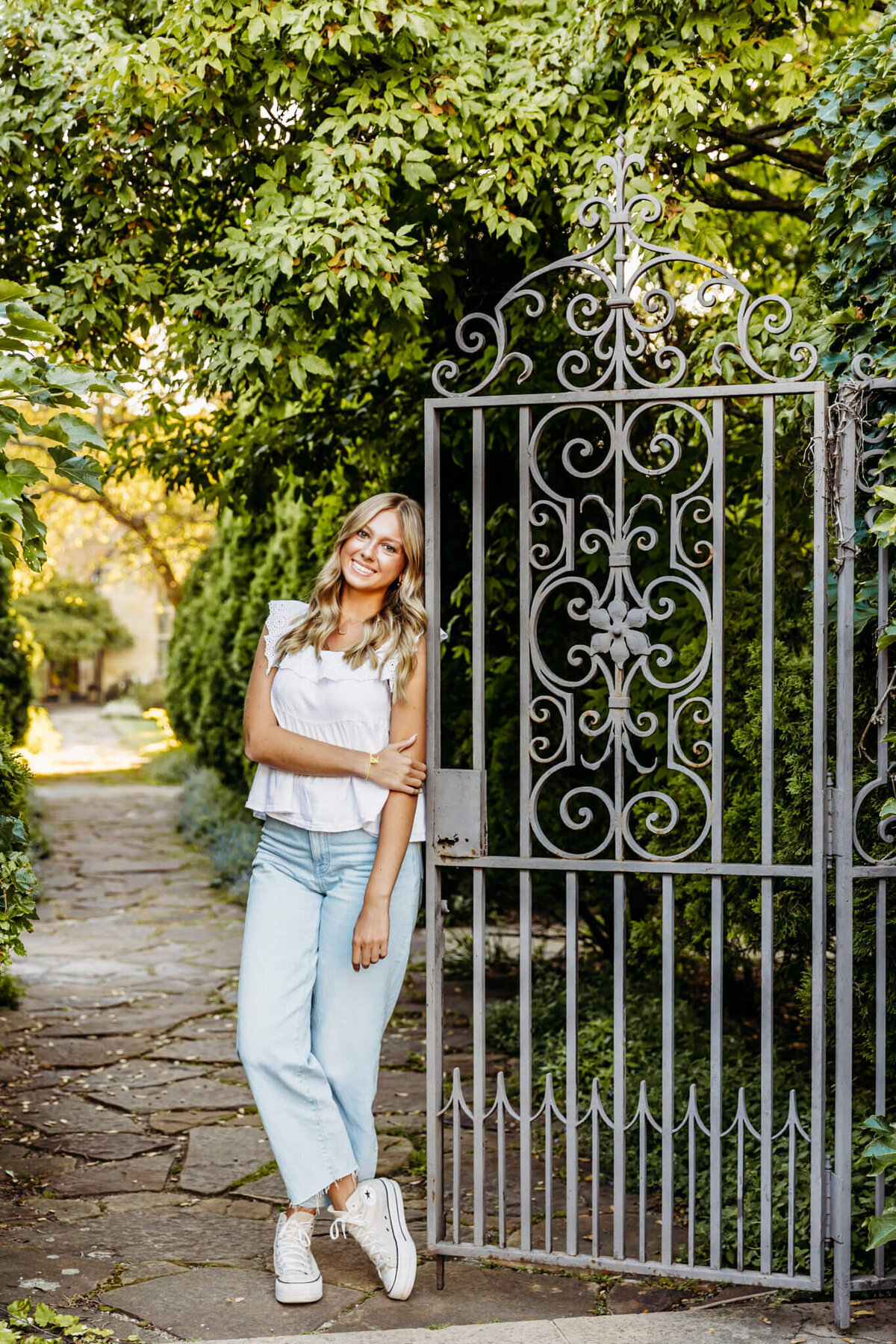 high school girl in a white top and jeans leaning against a garden gate while holding her left arm