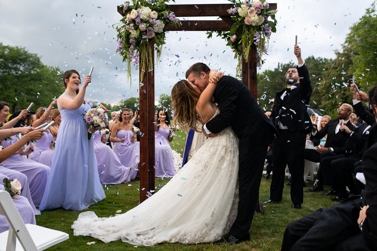 Bride and groom kiss at wedding ceremony as friends pop confetti to celebrate