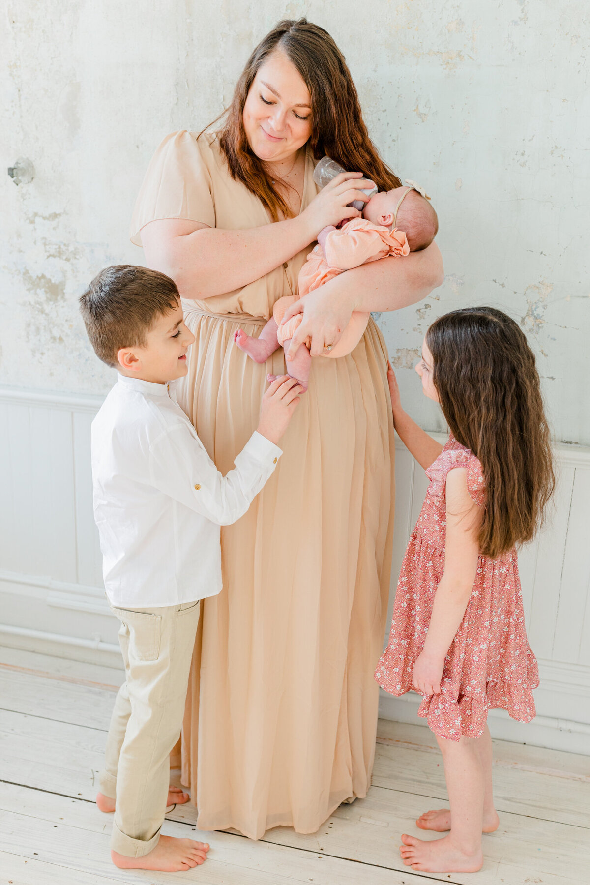 Newborn family session in Massachusetts | Mother is standing and feeding her newborn baby a bottle while brother plays with the baby's feet and sister looks on