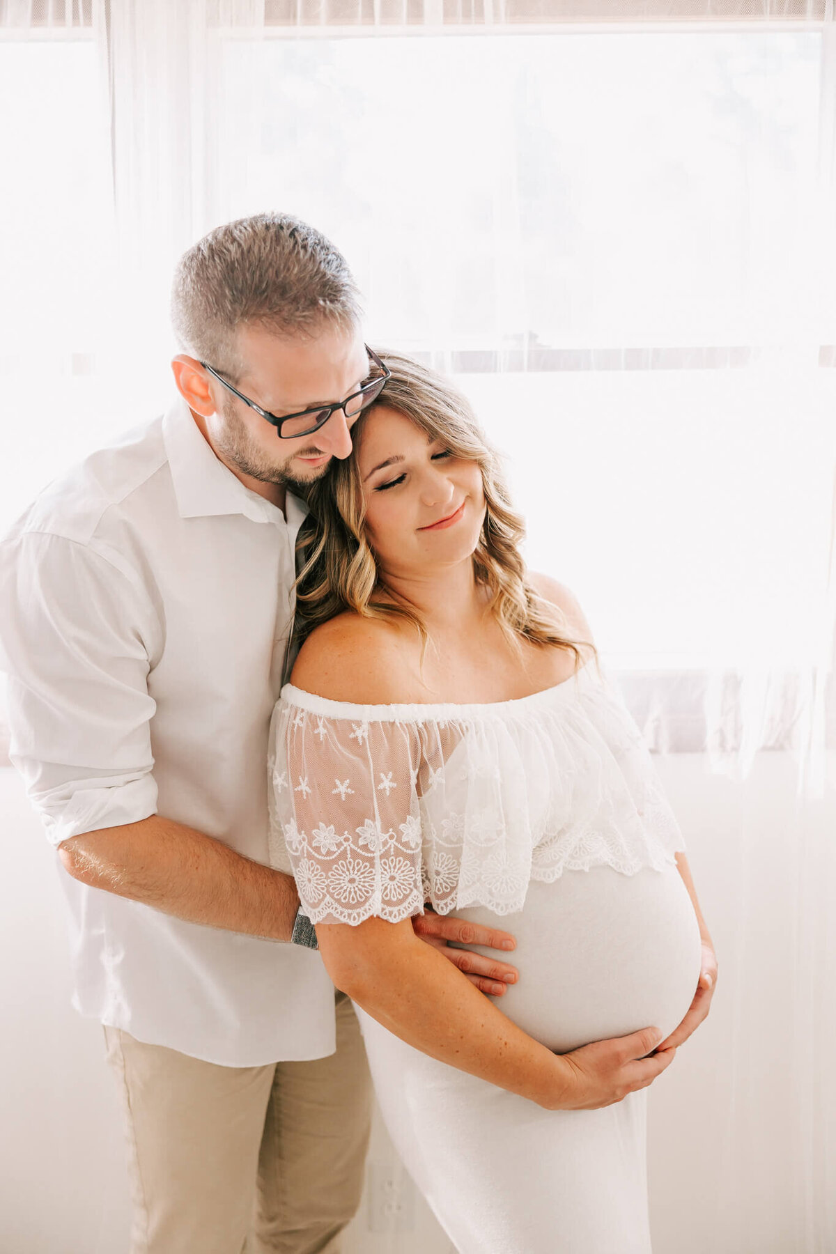 wife is wearing a white dress and leaning on husband  in front of studio window for her maternity session portraits in portland oregon.