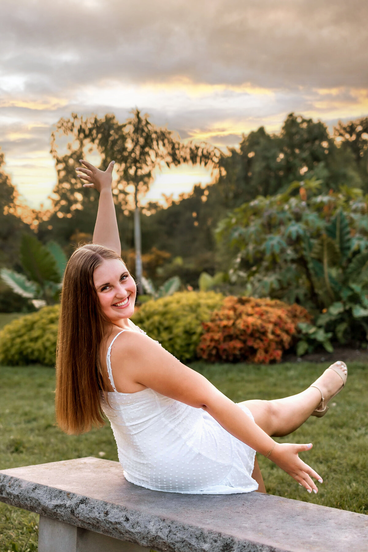 A brunette girl is having fun leaned back on a bench in tower grove park with her arms held open and her foot kicked up while she is looking back over her shoulder at her senior portrait photographer.