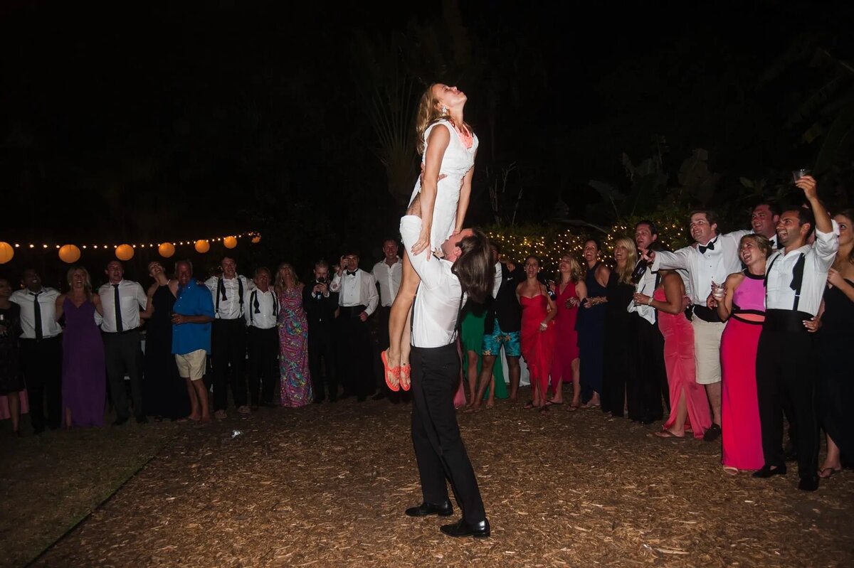 A bride is playfully lifted into the air during the wedding reception, surrounded by a cheering crowd under a night sky.