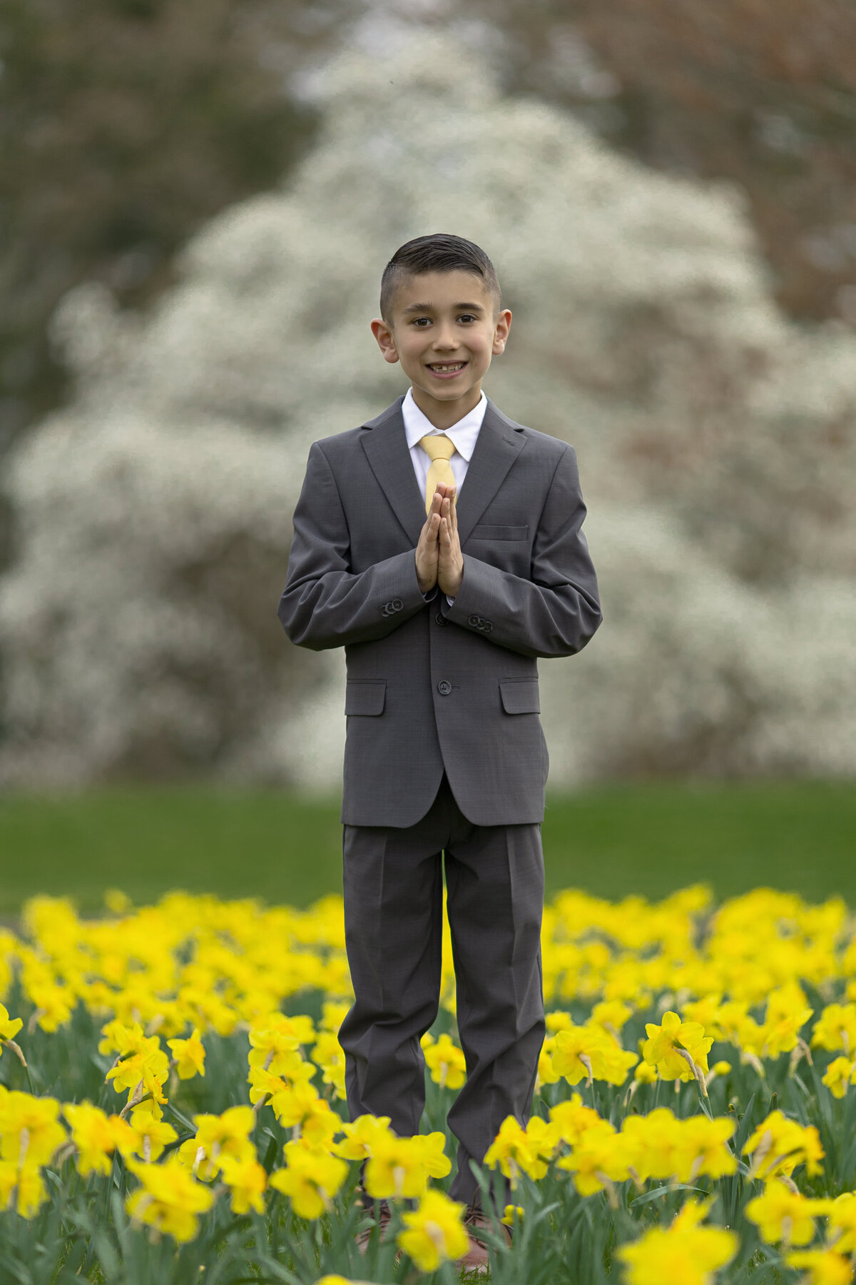 A boy prays with a smile while standing in a grey suit in a field of yellow daffodils