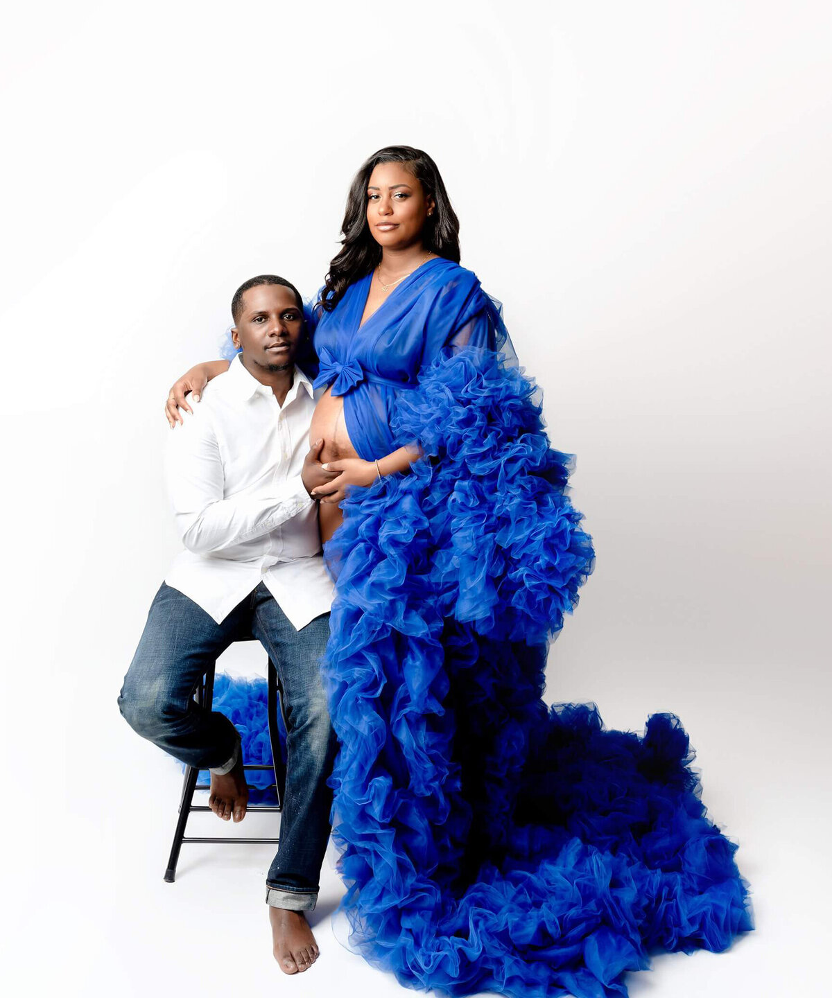 Husband and wife embrace during a maternity session. Woman wearing flowing dress with husband sitting beside her with hands on her belly.