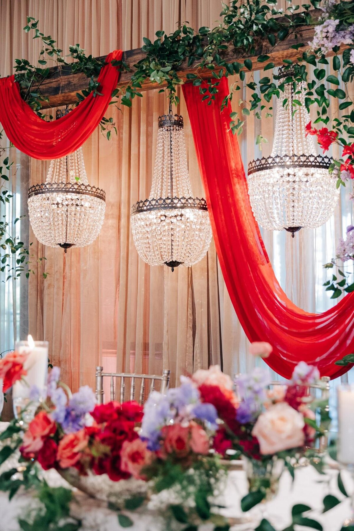 Elegant wedding reception decor with twin crystal chandeliers, draped red fabric, and a floral garland.