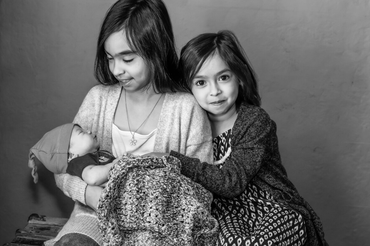 Indoor family lifestyle session with two girls and a newborn baby boy  in black and white.  Older sister is looking at the baby while younger sister is looking at the camera