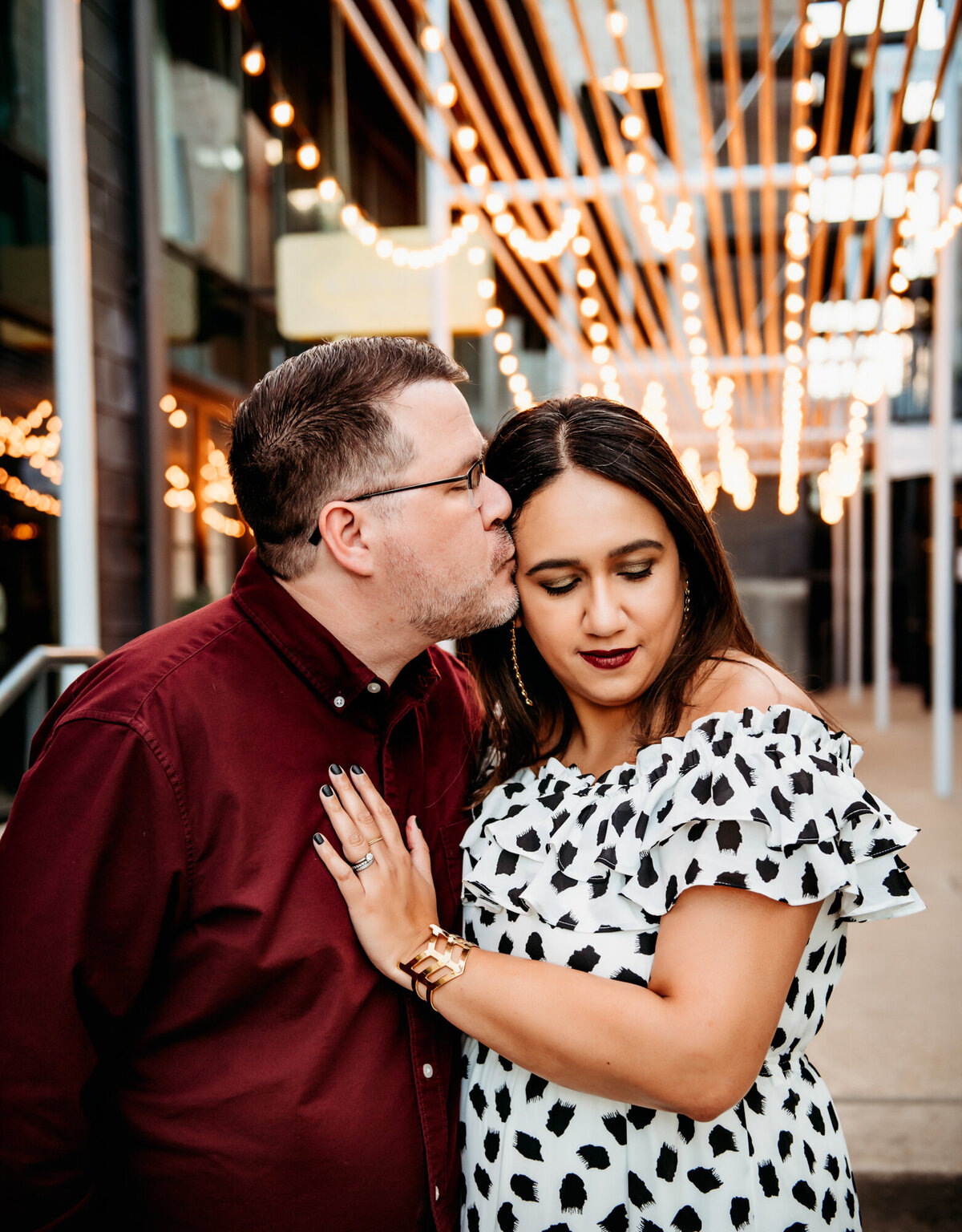 Couples Photography, Man in a maroon shirt kisses woman in black and white dress in front of the lights by Perla's