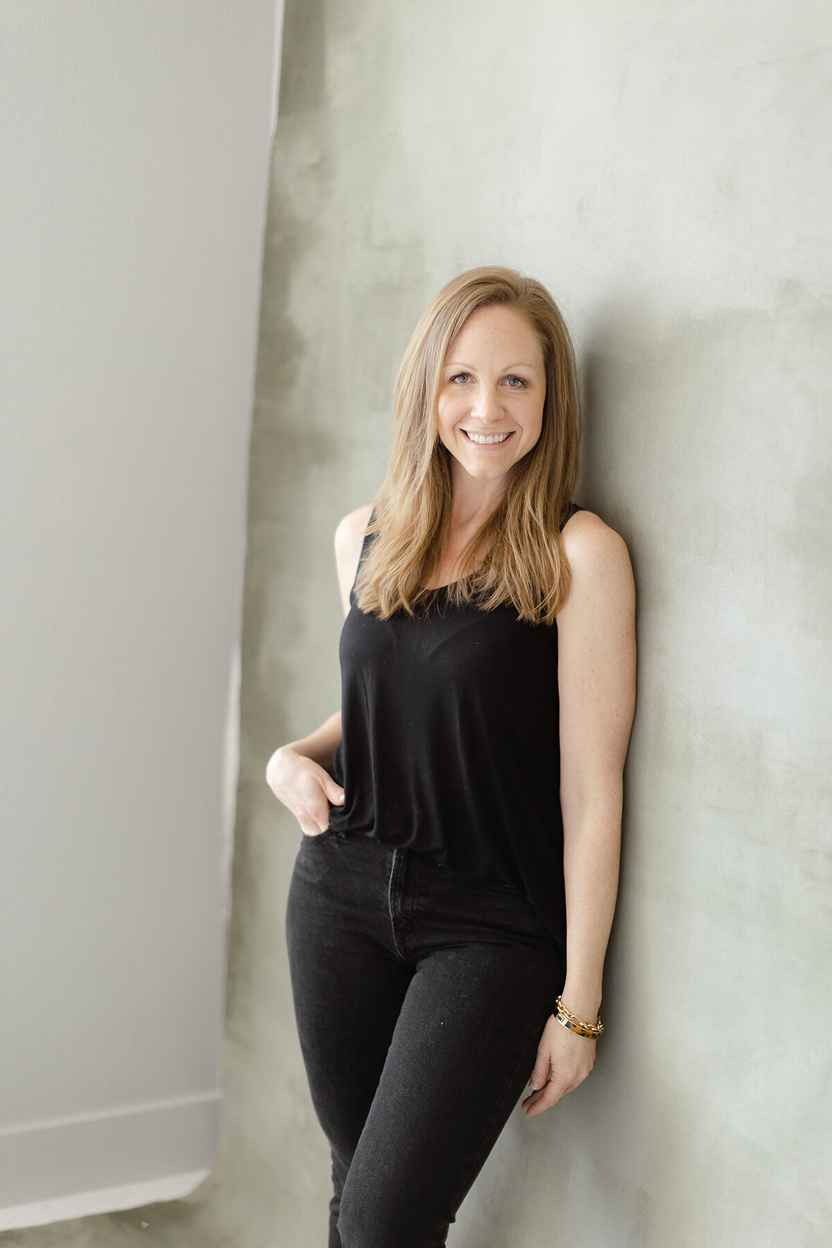 Professional headshot photo of a DFW business woman leaning against the wall in a Dallas photography studio as she has one hand in her pocket looking at the photographer smiling.