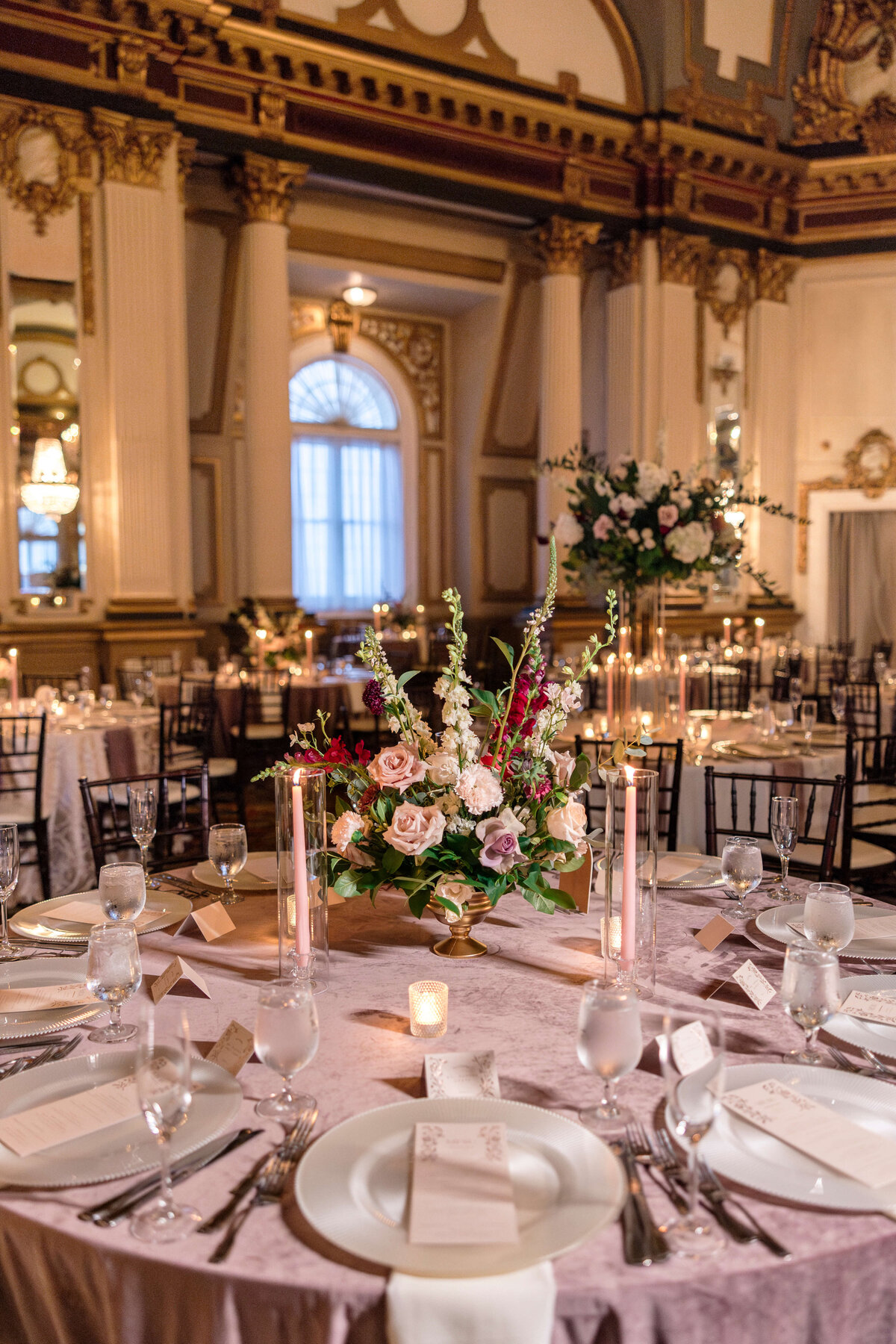 Elegant ballroom with ornate decor, featuring round tables set with white tablecloths, glassware, and floral centerpieces. Soft lighting enhances the luxurious atmosphere, with candles adding a warm glow.