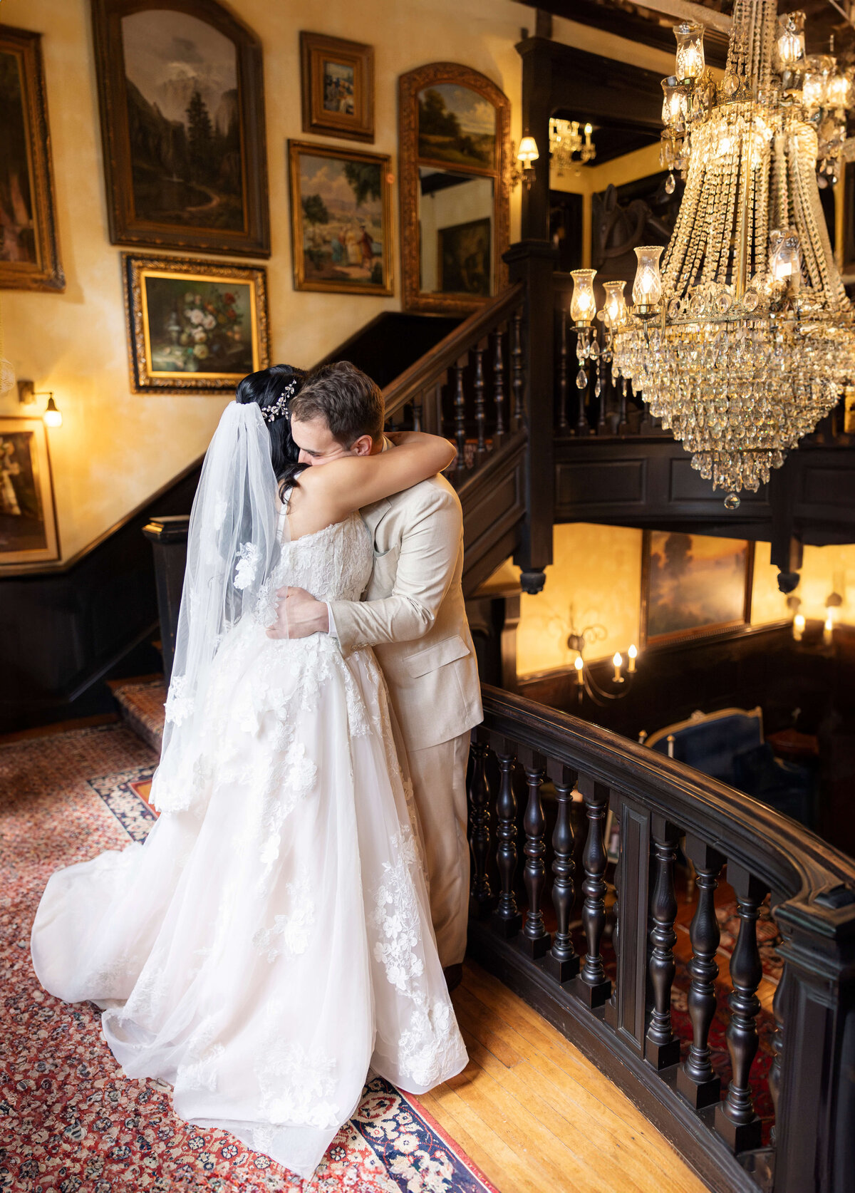 A couple embraces on a vintage staircase under a grand chandelier. The bride wears a lace gown and veil, while the groom is in a light-colored suit. The setting is adorned with framed paintings and warm lighting.