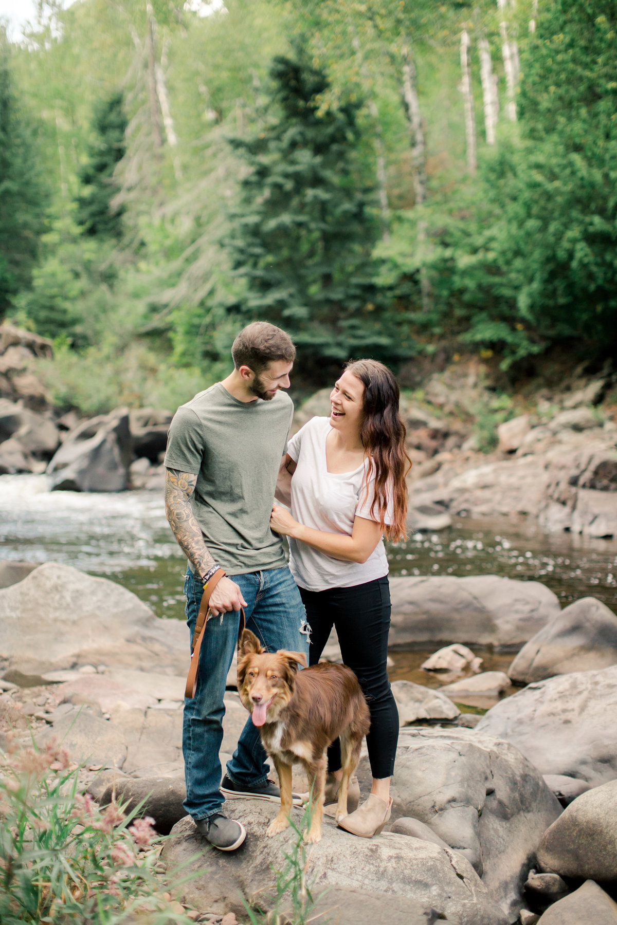 couple laughs together with dog for portraits beside river in temperance river state park