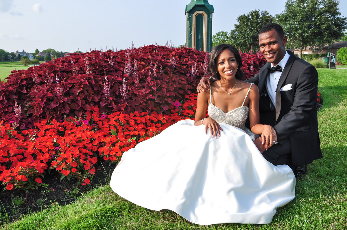 Bride sitting down outside and groom standing, behind them are beautiful florals and tall clock