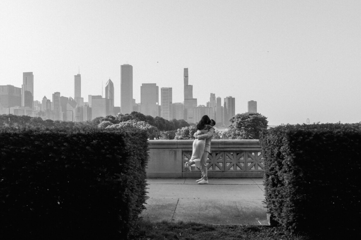 A black and white engagement photo of a couple embracing in front of the city skyline
