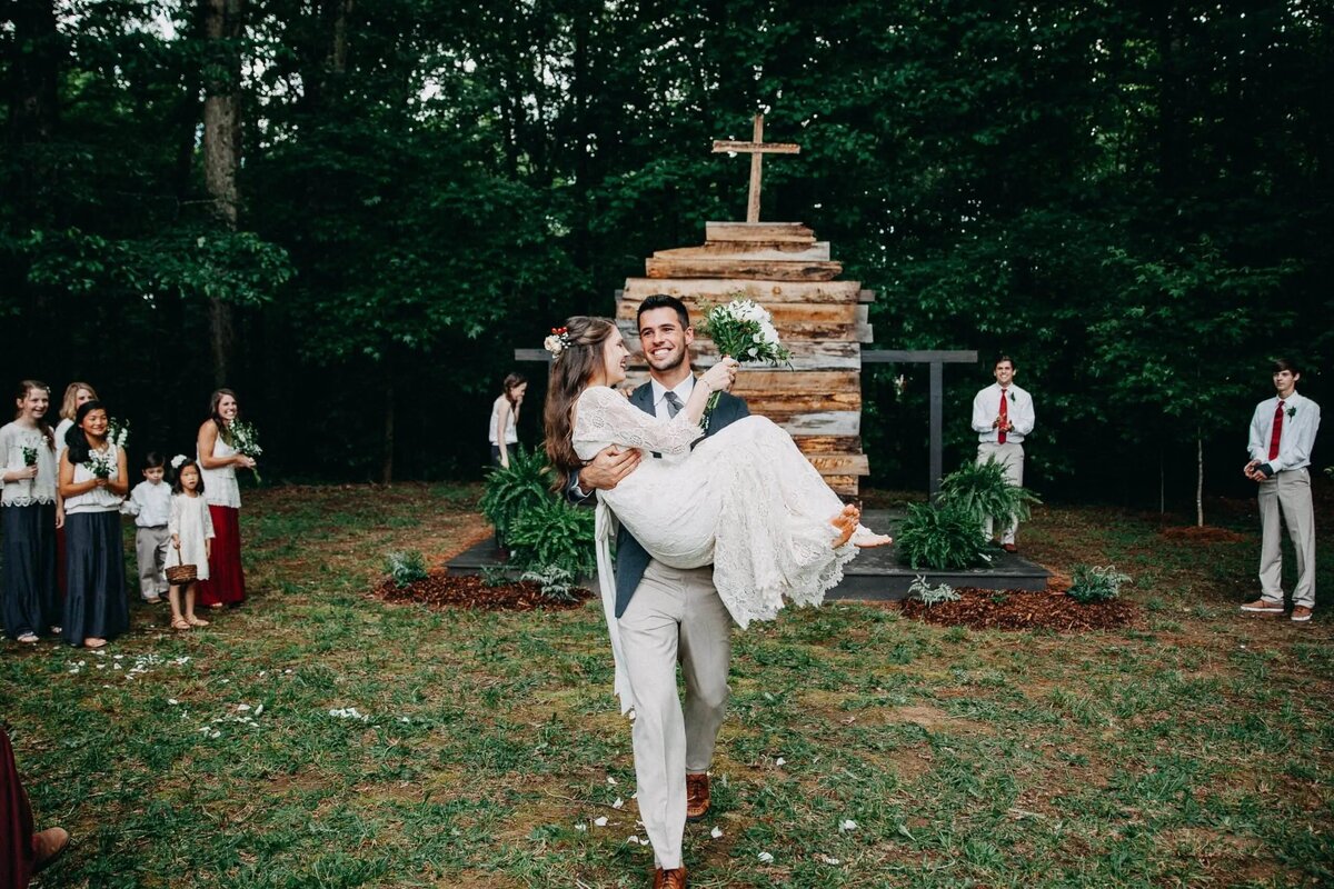 A groom carrying his bride in front of a wooden cross during their outdoor wedding, symbolizing a joyous and traditional ceremony.