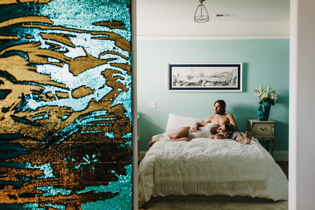 In home intimate maternity session with pregnant mom lying on bed and dad sitting up on bed. Both gazing toward window with gold and turquoise sequin art on wall