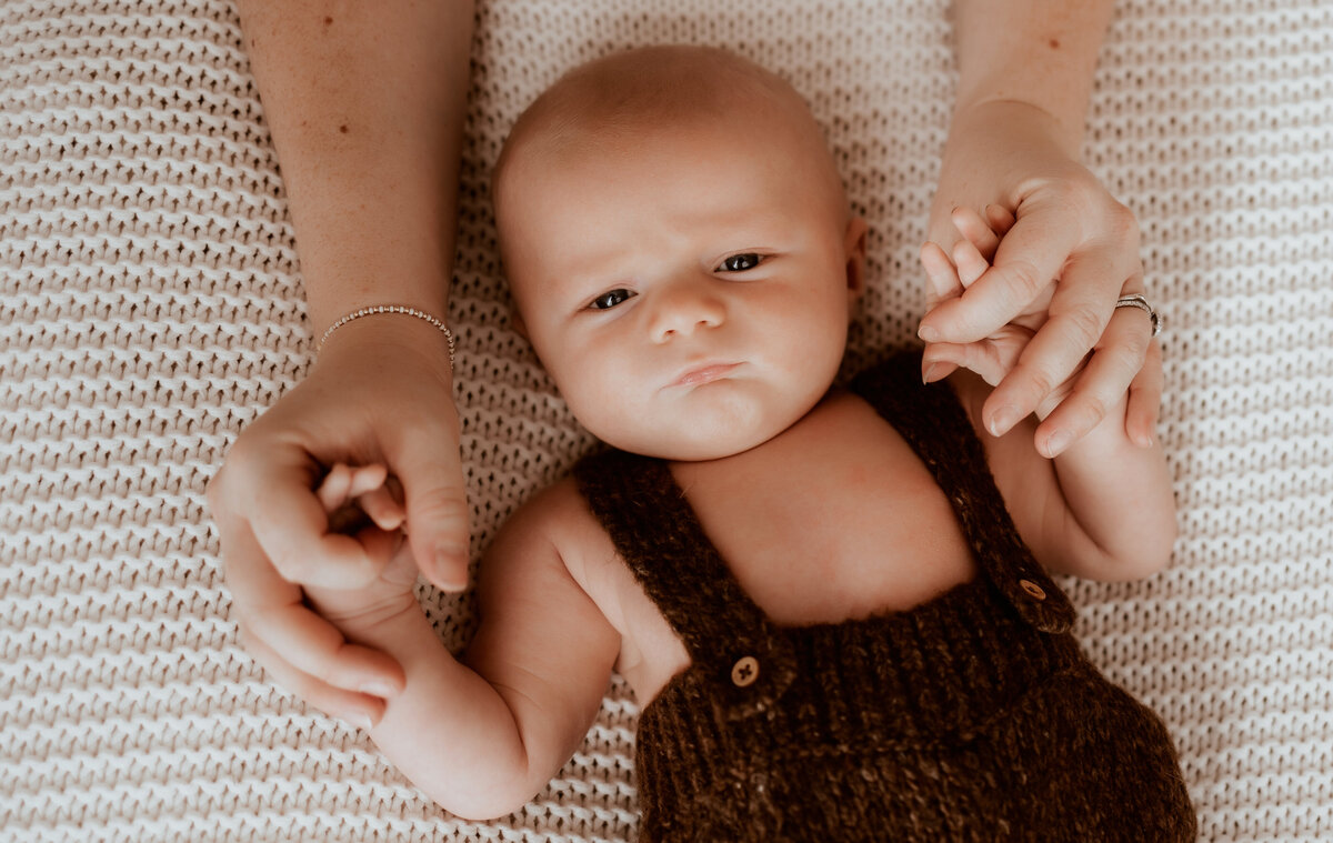 baby boy holds moms hands wearing  brown knit overalls