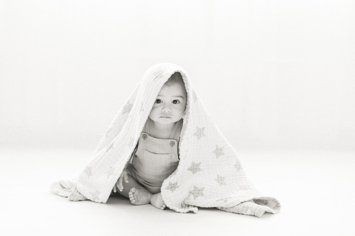 Black and white photo of a baby boy playing under a blanket and looking at the Fort Worth photographer.
