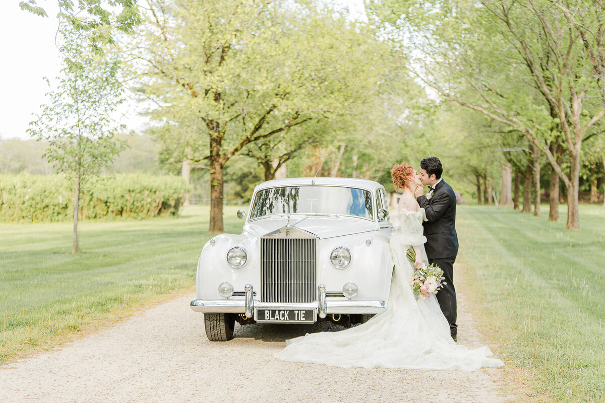 A bride and groom share a kiss on an estate in Boston's North Shore. The bride is leaning against a vintage Rolls Royce. Captured by best Massachusetts wedding photographer Lia Rose Weddings
