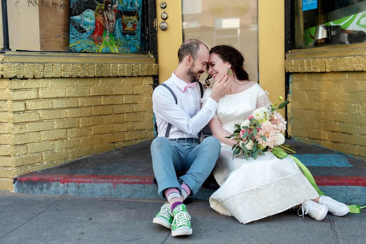 a bride and groom sit on a stoop hugging together.  The bride holds a pretty ivory and cream bouquet by flowers by alana