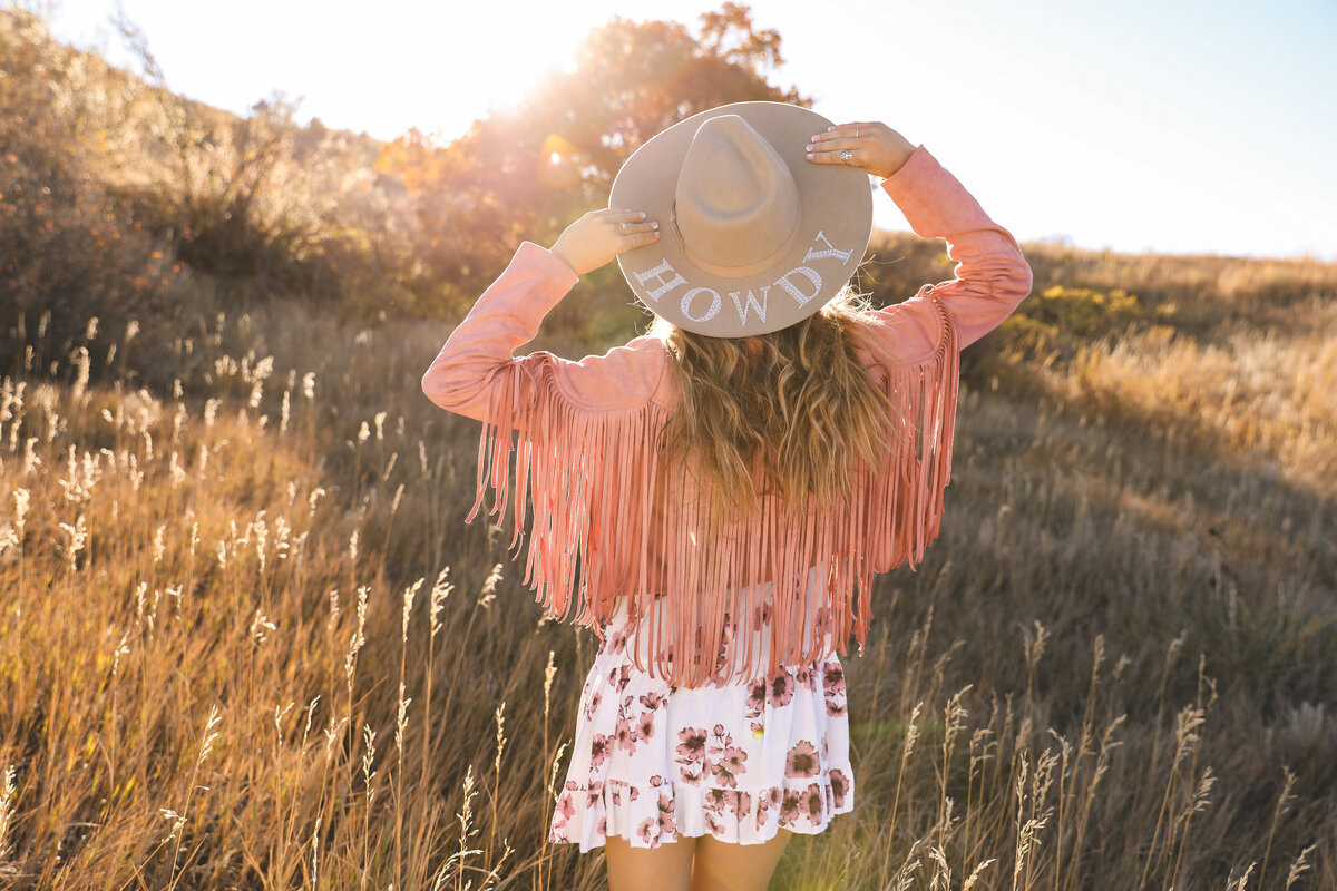 Sun-filled styled senior picture of a girl in a western-inspired outfit wearing a Howdy cowboy hat