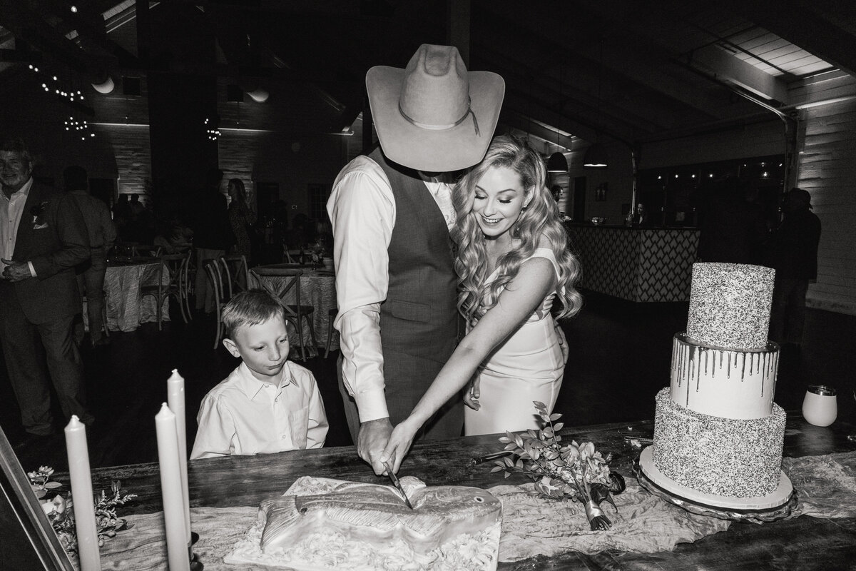 A black and white photograph of Carly and Bradley cutting their cake on their wedding day at Morgan Creek Barn in Dripping Springs, near Austin, Texas. The bride and groom are facing us as they cut their cake, in the shape of a fish. There is a three tiered wedding cake to their left covered in sprinkles and a child to their right watching them cut into the fish-shaped cake. Wedding photography taken by Stacie McChesney/Vitae Weddings.
