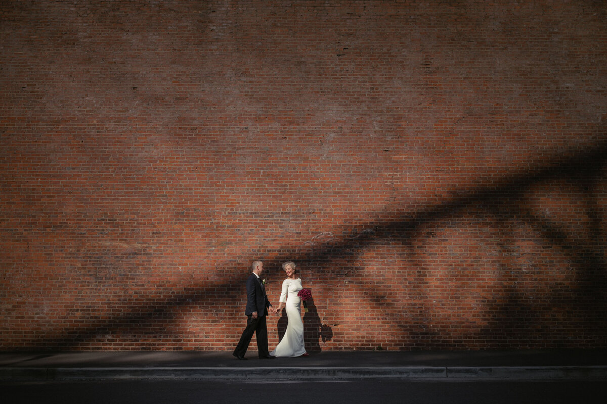 A wedding couple holding hands and walking along the side of a red brick building.