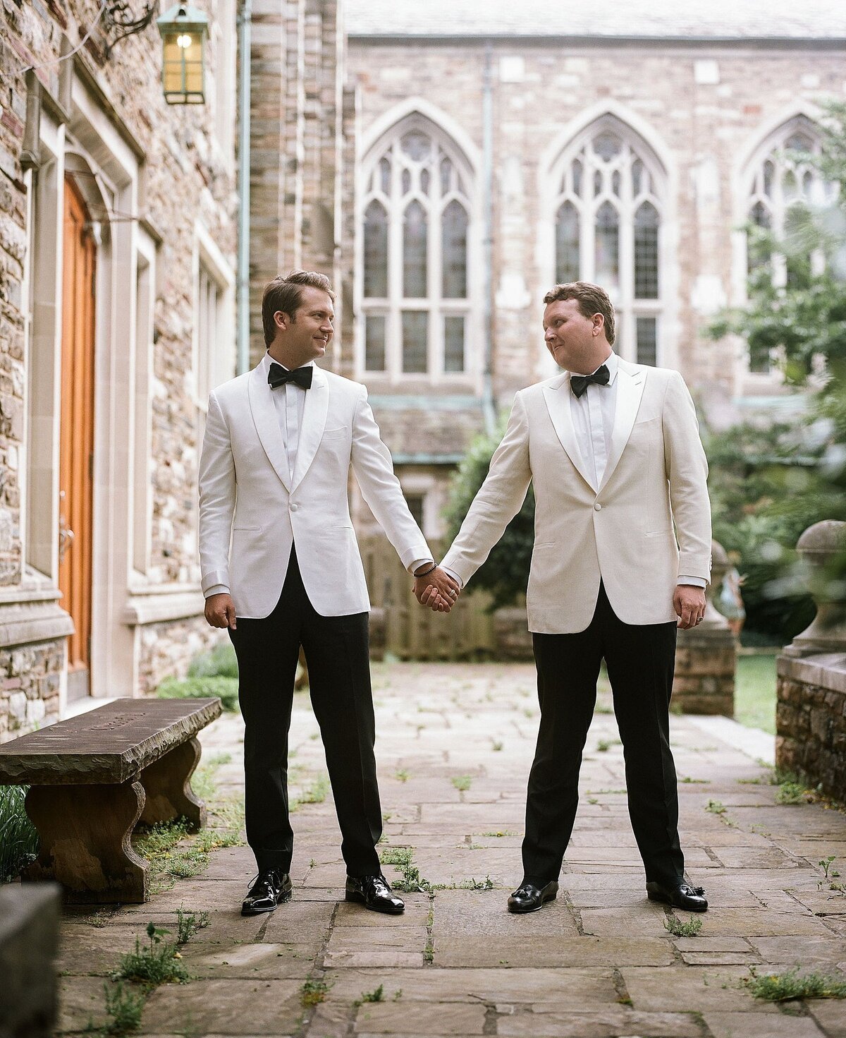 Two grooms wearing tuxedos with white jackets and black pants holding hands on a cobblestone path at Scarritt Bennett Center in Nashville for their LGBTQIA wedding